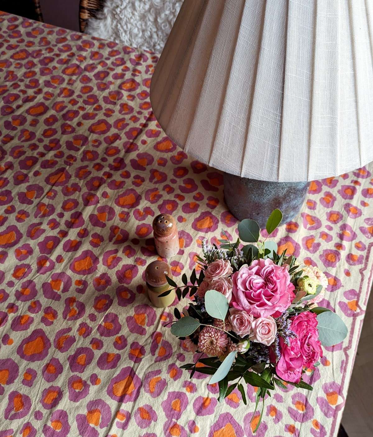 Eine Tischdecke mit Leopardenmuster in Pink und Orange liegt auf einem Tisch. Auf der Tischdecke stehen Salz- und Pfefferstreuer aus Keramik. Außerdem ein kleiner Blumenstrauß und eine Tischlampe.
